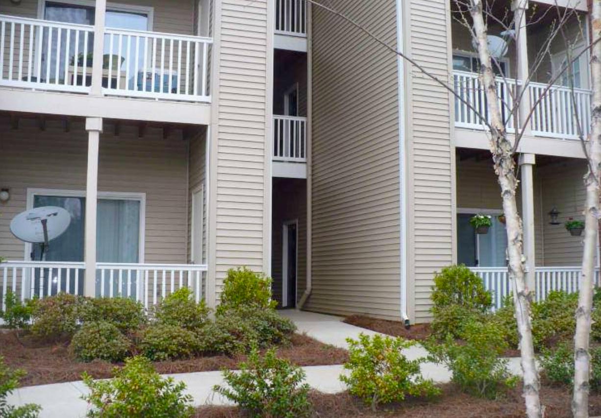 Beige apartment building with balconies, white railings, shrubs, and a satellite dish outside.