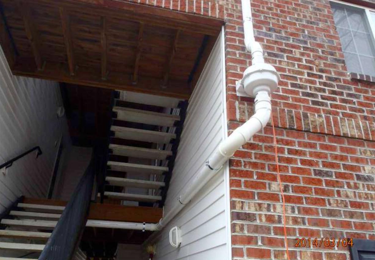 White PVC pipes run vertically and horizontally along a brick and white-paneled building near exterior stairs.