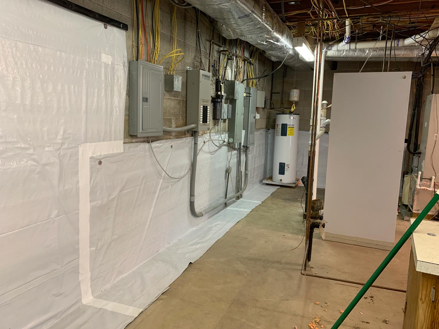 A basement with exposed pipes, electrical panels, a water heater, and plastic wall and floor covering.