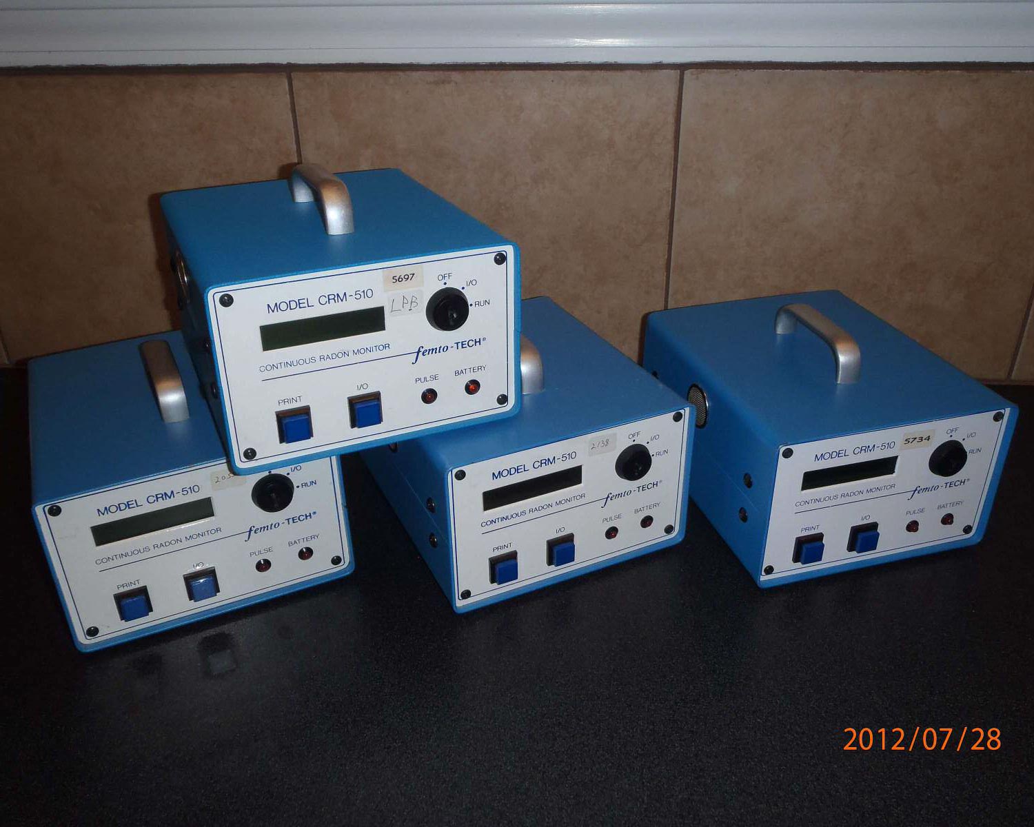 Four blue electronic radon monitors labeled "FAMILY TECH" on a countertop against a tiled wall.