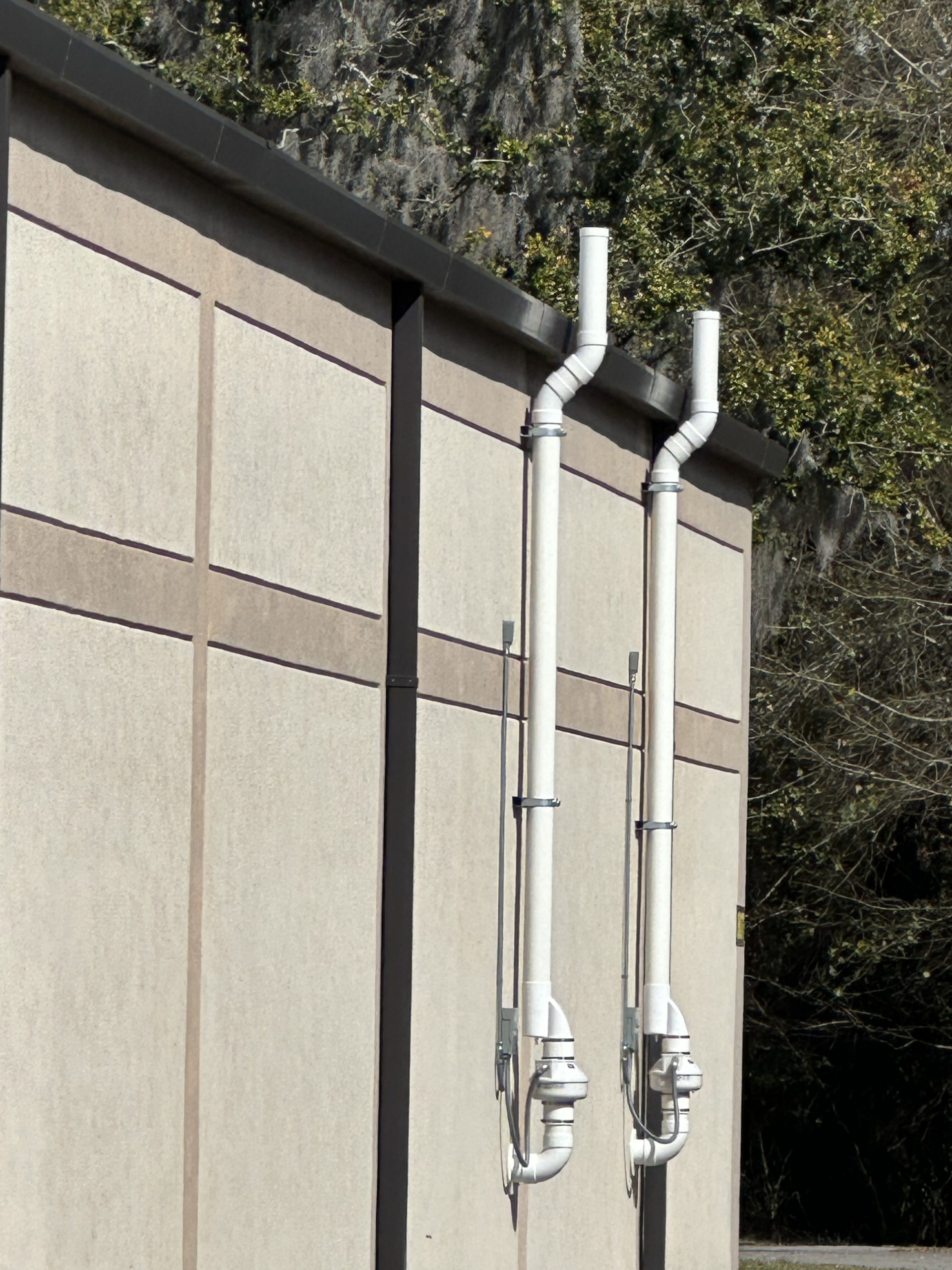 Two white pipes run vertically up the side of a beige building with trees in the background.
