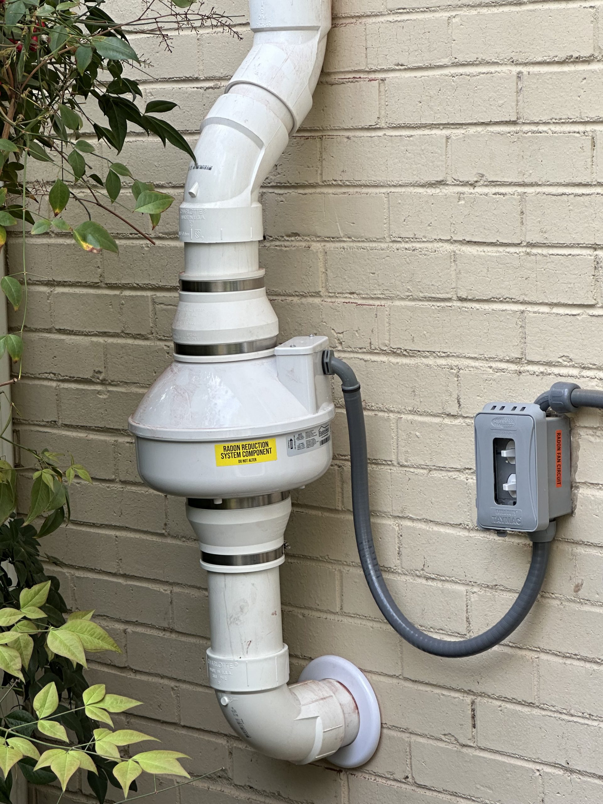 White radon mitigation system with pipes attached to a brick wall, next to electrical components and green plants.