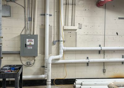 Pipes and conduits run along a basement wall; tools and PVC pipes are scattered on the floor.