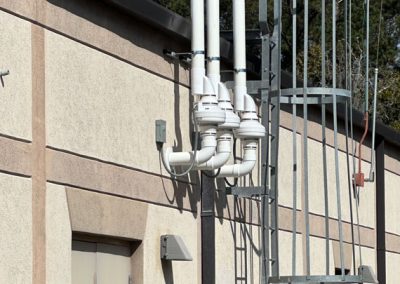 Four vertical white pipes with curved bases are mounted on the exterior wall of a beige building near a metal ladder.
