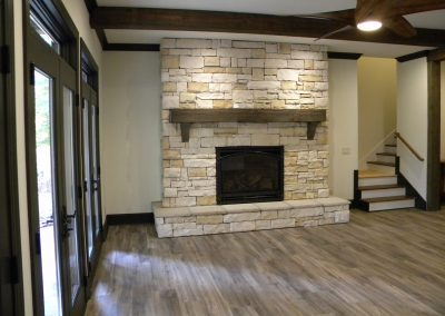 Stone fireplace with wooden mantel in a modern living room with wood floors and stairs nearby.
