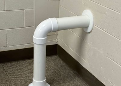 A white PVC pipe bends from the wall and floor, resembling a question mark in a carpeted corner.