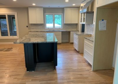 Modern kitchen with white cabinets, marble countertops, an island, and hardwood floors, viewed in daylight.