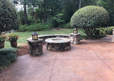 A stone fire pit with lanterns on a patio, surrounded by bushes and potted plants in a garden setting.