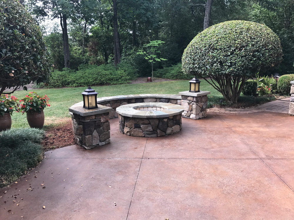 Stone fire pit with lanterns on a patio, surrounded by trimmed bushes and potted plants in a garden.
