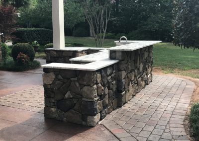 Outdoor stone bar with a marble countertop and sink, set on a paved patio with greenery in the background.