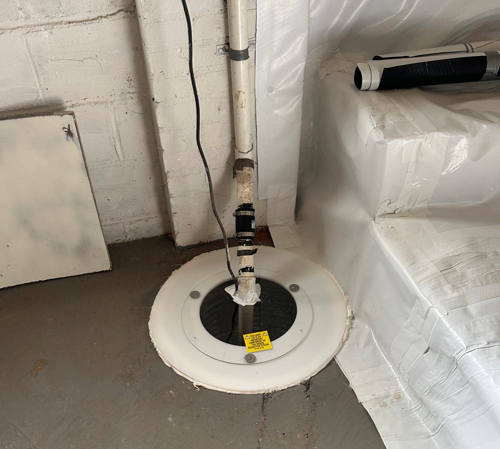 A sump pump system installed in a basement floor near a wall, wrapped pipes connected above it.