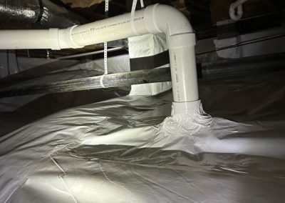 White PVC pipes run through a dimly lit crawl space with a plastic vapor barrier covering the floor.