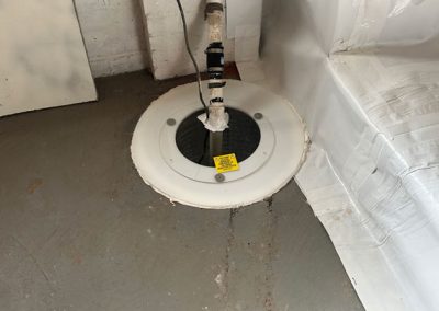 A sump pump cover with a warning label is installed on a gray basement floor near a wall and plastic-covered stairs.