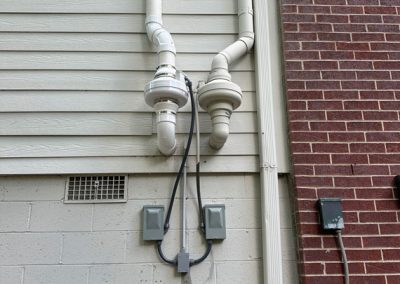 Two white ventilation pipes with caps run up the side of a building next to a brick wall and utility boxes.